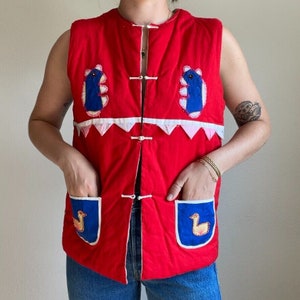 May include: Red quilted vest with blue and white embroidered birds and geometric designs. The vest has a button closure and two pockets.