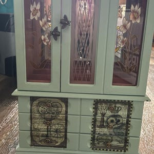 May include: A light green jewelry armoire with glass doors and decorative floral accents. The doors have bee-shaped clasps. The bottom features two drawers with vintage-style illustrations. The armoire is on a wooden surface.