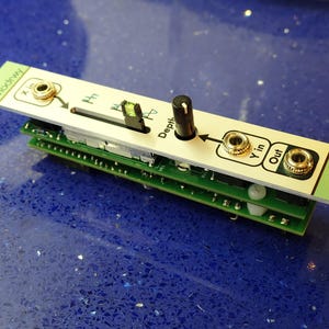 K&ouml;nnte beinhalten: Ein Modul f&uuml;r einen modularen Synthesizer mit einer wei&szlig;en Frontplatte und einer gr&uuml;nen Leiterplatte. Die Frontplatte hat einen schwarzen Knopf, einen Schieberegler und goldfarbene Buchsen mit der Aufschrift "X in", "Y in" und "Out". Das Modul ist auf anderen gr&uuml;nen Leiterplatten gestapelt.