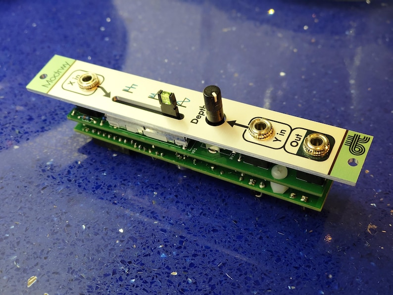 K&ouml;nnte beinhalten: Ein Modul f&uuml;r einen modularen Synthesizer mit einer wei&szlig;en Frontplatte und einer gr&uuml;nen Leiterplatte. Die Frontplatte hat einen schwarzen Knopf, einen Schieberegler und goldfarbene Buchsen mit der Aufschrift "X in", "Y in" und "Out". Das Modul ist auf anderen gr&uuml;nen Leiterplatten gestapelt.