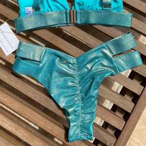 May include: Teal and gold glitter bikini top and bottom. The top has a halter style with adjustable straps and a clasp closure in the back. The bottom has a high-cut style with a gold glitter trim and cutouts on the sides.