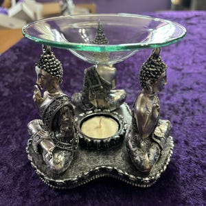 May include: Silver-toned metal incense burner with three seated Buddha figures and a glass dish for essential oils. The burner has a decorative base with a floral design.