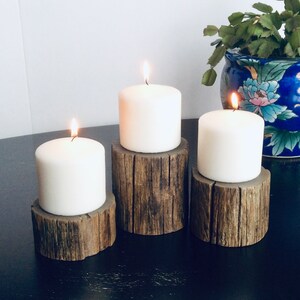 Peut inclure: Trois bougies blanches en forme de pilier sur des bougeoirs en bois rustique. Les bougies sont allum&eacute;es et les bougeoirs sont en bois naturel avec une texture rugueuse.
