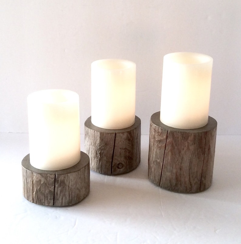 Peut inclure: Trois bougies blanches en forme de pilier sur des bases en bois. Les bases sont faites pour ressembler &agrave; des souches d'arbres. Les bougies sont allum&eacute;es et brillent.