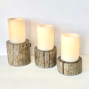 Peut inclure: Trois bougies blanches en forme de pilier sur des bases en bois rustique. Les bougies sont allum&eacute;es et brillent.