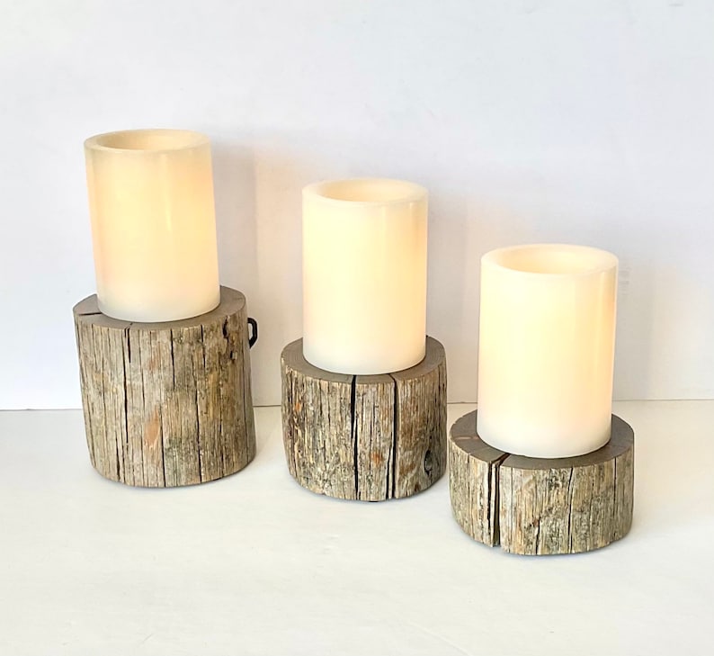Peut inclure: Trois bougies blanches en forme de pilier sur des bases en bois rustique. Les bougies sont allum&eacute;es et brillent.