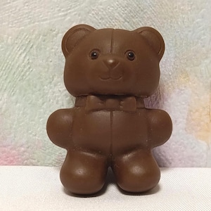 May include: A solid brown teddy bear figurine with a bow tie. The bear has a classic design with rounded features, including a small nose and button eyes. The toy is made of a smooth material and stands upright.
