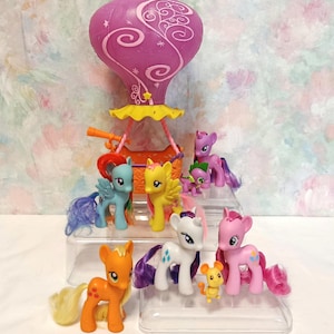 Puede incluir: Un juego de My Little Pony con varias figuras de pony coloridas y una gran estructura en forma de linterna púrpura. Los ponis son de varios colores, incluyendo rosa, amarillo, azul y naranja. El juego se exhibe en una plataforma transparente de varios niveles.