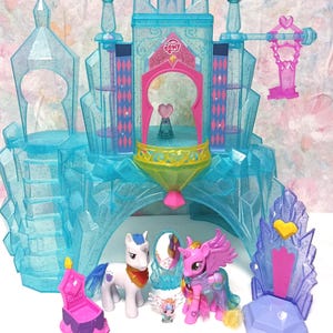 Könnte beinhalten: Ein durchscheinendes blaues Spielzeugschloss aus Kunststoff mit Glitzerakzenten, einem zentralen Turm, einem rosa Torbogen und dem My Little Pony-Logo. Enthalten sind zwei Ponyfiguren, eine kleine Kreatur, ein rosa Stuhl, ein Spiegel und ein violetter Thron.