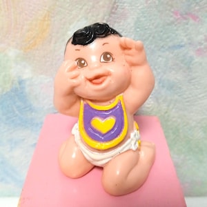 May include: A vintage baby figurine with black hair, rosy cheeks, and a smiling expression. The baby is wearing a purple and yellow bib with a heart design and a white diaper. The figurine is kneeling on a pink surface.
