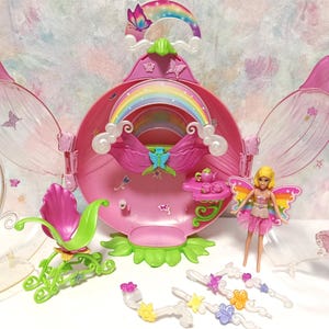 以下が含まれることがあります： ピンクと緑の花の形をしたドールハウスのプレイセット。半透明の花びらのドア、虹のアーチ、ティーセット付きの小さなテーブルが特徴です。虹色の羽を持つブロンドの人形、緑のロッキングチェア、4つの透明なプラスチック製のつるのアクセサリーが含まれています。