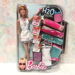Puede incluir: Un set de muñeca Barbie H2O Design Studio en su empaque original. La muñeca rubia viste un vestido blanco con detalles plateados. Incluye un marcador rosa, un aplicador magenta, una herramienta turquesa, un vestido y muestras de tela.