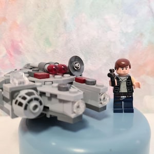 Op de afbeelding: Een Lego Millennium Falcon model met een Lego figuur van Han Solo. Het ruimteschip is grijs met rode en transparante accenten. De Han Solo figuur draagt een zwart vest en houdt een zwarte blaster vast.
