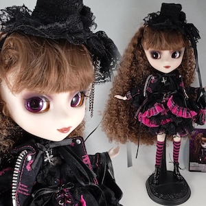 Könnte beinhalten: Modepuppe mit braunem Lockenhaar, in einem schwarzen Outfit mit rosa Akzenten, passendem Hut und Stiefeln. Die Puppe hat große violette Augen und wird mit einer Miniaturversion präsentiert.