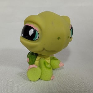 May include: A green plastic turtle toy with pink accents and large blue eyes. The turtle is sitting on its hind legs with its front legs outstretched.