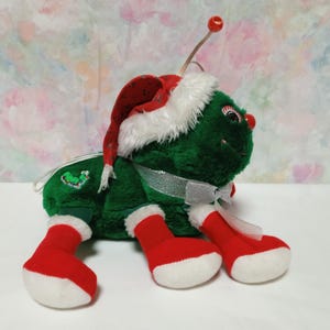 K&ouml;nnte beinhalten: Ein gr&uuml;nes Pl&uuml;schtier in Form eines K&auml;fers, das einen roten und wei&szlig;en Weihnachtsmann-Hut und wei&szlig;e Stiefel tr&auml;gt. Der K&auml;fer hat ein wei&szlig;es Band um den Hals und ein gr&uuml;nes und rotes Stechpalmen-Design auf dem R&uuml;cken.