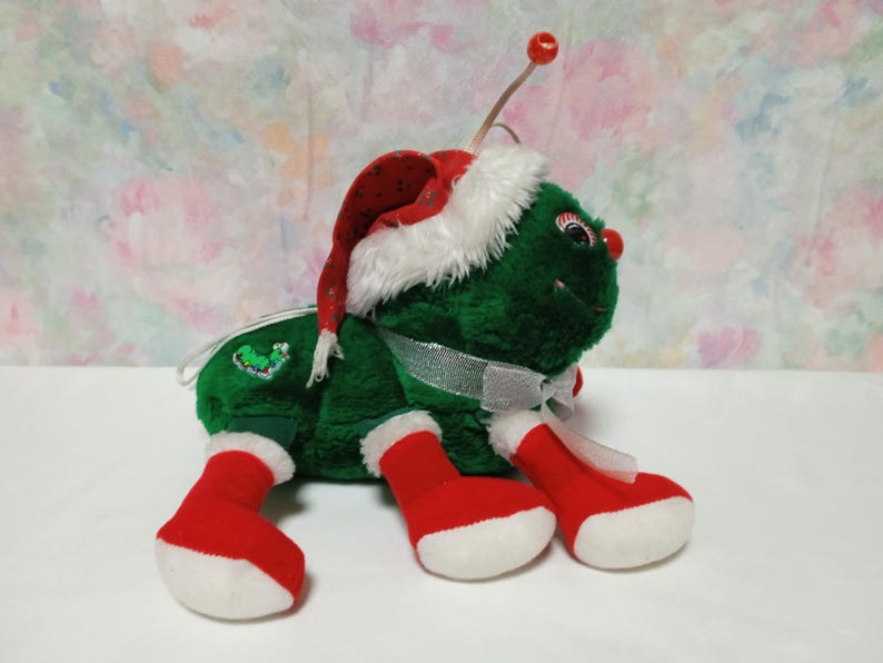 K&ouml;nnte beinhalten: Ein gr&uuml;nes Pl&uuml;schtier in Form eines K&auml;fers, das einen roten und wei&szlig;en Weihnachtsmann-Hut und wei&szlig;e Stiefel tr&auml;gt. Der K&auml;fer hat ein wei&szlig;es Band um den Hals und ein gr&uuml;nes und rotes Stechpalmen-Design auf dem R&uuml;cken.