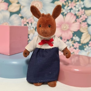 May include: A brown plush toy with a white shirt and a red bow tie. The toy is wearing a blue skirt and has a deer-like face.
