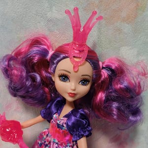 K&ouml;nnte beinhalten: Puppe mit rosa und lila Haaren in Z&ouml;pfen, die einen rosa Kragen und ein lila und gebl&uuml;mtes Kleid tr&auml;gt. Die Puppe hat blaue Augen und roten Lippenstift. Die Puppe ist ein Spielzeug.