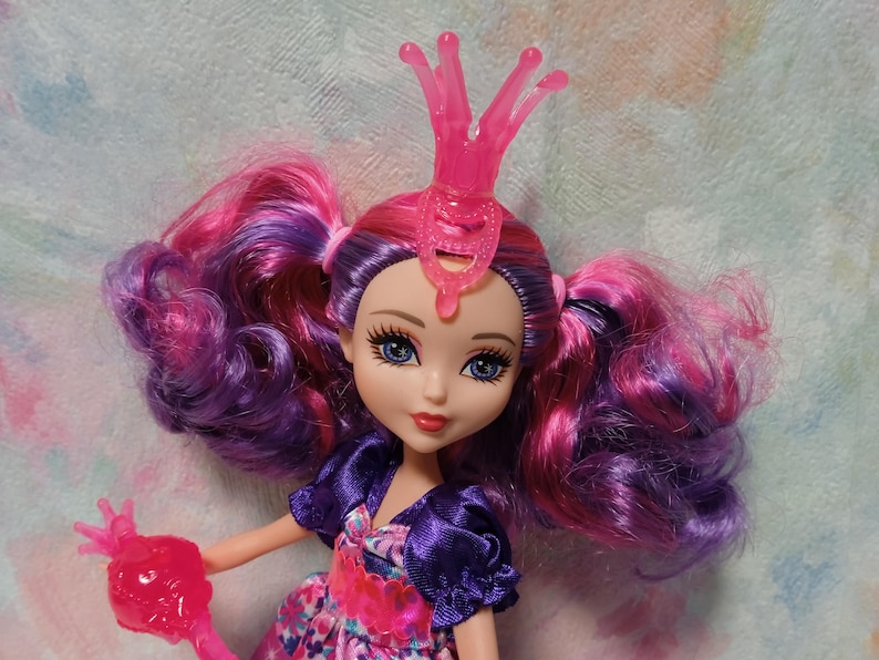 K&ouml;nnte beinhalten: Puppe mit rosa und lila Haaren in Z&ouml;pfen, die einen rosa Kragen und ein lila und gebl&uuml;mtes Kleid tr&auml;gt. Die Puppe hat blaue Augen und roten Lippenstift. Die Puppe ist ein Spielzeug.