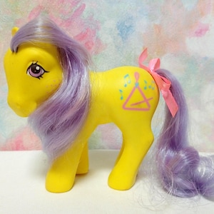 May include: A yellow toy pony with a purple mane and tail, and a pink ribbon tied to the tail. The pony has a musical triangle design on its side. The background is a soft, colorful pattern.