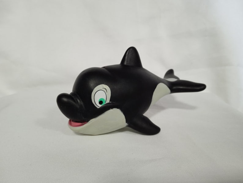 K&ouml;nnte beinhalten: Schwarz-wei&szlig;es Spielzeug-Orca aus Gummi mit einem gr&uuml;nen Auge und einem wei&szlig;en Bauch. Der Orca ist in Schwimmposition mit offenem Maul.