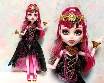 Peut inclure: Une poupée avec des cheveux roses et une couronne, des boucles d'oreilles et des bracelets dorés. La poupée porte une robe noire avec des détails roses et un corsage doré. La poupée a des chaussures roses et un collier doré. Une poupée de collection.