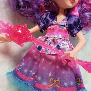 K&ouml;nnte beinhalten: Eine Puppe mit lila Haaren und einem rosa und lila Kleid. Die Puppe h&auml;lt einen rosa, durchscheinenden Zauberstab. Das Kleid hat ein florales Oberteil und einen Rock mit Sternenmuster. Die &Auml;rmel der Puppe sind dunkelviolett.