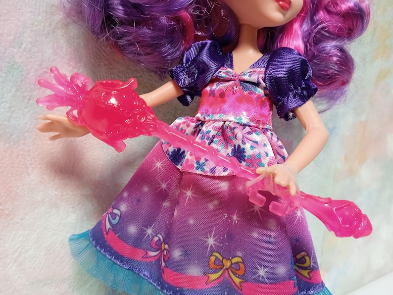 K&ouml;nnte beinhalten: Eine Puppe mit lila Haaren und einem rosa und lila Kleid. Die Puppe h&auml;lt einen rosa, durchscheinenden Zauberstab. Das Kleid hat ein florales Oberteil und einen Rock mit Sternenmuster. Die &Auml;rmel der Puppe sind dunkelviolett.
