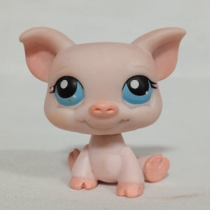 Puede incluir: Un juguete de plástico rosa en forma de cerdo con ojos azules y una nariz rosa. El cerdo está sentado sobre una superficie blanca.