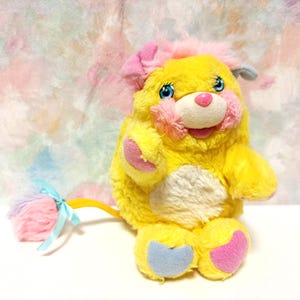 May include: A vintage yellow plush toy with pink and blue accents. The toy has a fluffy yellow body, pink ears, and a pink nose. It has blue eyes and a white belly. A colorful tail with a blue ribbon is attached to the toy.
