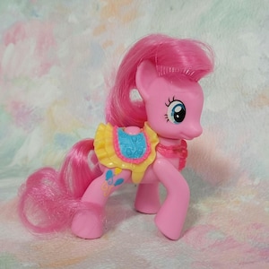 May include: Pink My Little Pony toy with a yellow, blue, and pink patterned dress. The pony has pink hair and a pink body.
