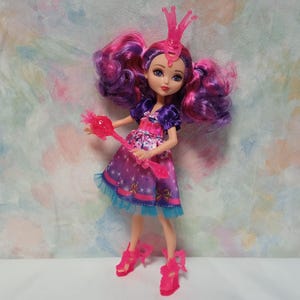 K&ouml;nnte beinhalten: Puppe mit rosa und lila Haaren, tr&auml;gt ein lila Kleid mit Sternenmuster und eine rosa Krone. Die Puppe h&auml;lt einen rosa Zauberstab und tr&auml;gt rosa Schuhe. Das Kleid der Puppe hat eine blaue Bord&uuml;re.