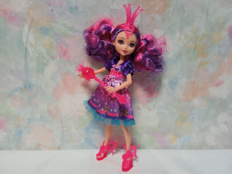 K&ouml;nnte beinhalten: Puppe mit rosa und lila Haaren, tr&auml;gt ein lila Kleid mit Sternenmuster und eine rosa Krone. Die Puppe h&auml;lt einen rosa Zauberstab und tr&auml;gt rosa Schuhe. Das Kleid der Puppe hat eine blaue Bord&uuml;re.