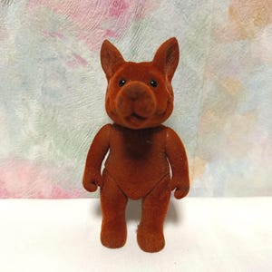 May include: A brown, plush dog figurine with upright ears and a rounded snout. The dog is standing upright on a white surface, against a backdrop of pastel colors. The toy is a solid color and has a soft, velvety texture.