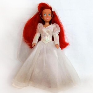K&ouml;nnte beinhalten: Eine Puppe mit roten Haaren und blauen Augen, die ein wei&szlig;es Kleid mit langen &Auml;rmeln und einem Schleier tr&auml;gt. Das Kleid der Puppe hat ein glitzerndes Oberteil und einen weiten Rock. Die Puppe steht aufrecht.