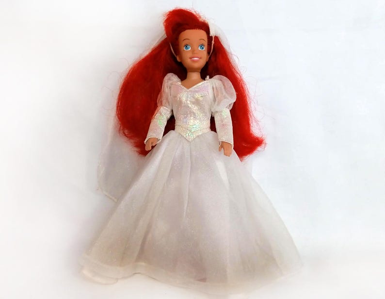 K&ouml;nnte beinhalten: Eine Puppe mit roten Haaren und blauen Augen, die ein wei&szlig;es Kleid mit langen &Auml;rmeln und einem Schleier tr&auml;gt. Das Kleid der Puppe hat ein glitzerndes Oberteil und einen weiten Rock. Die Puppe steht aufrecht.