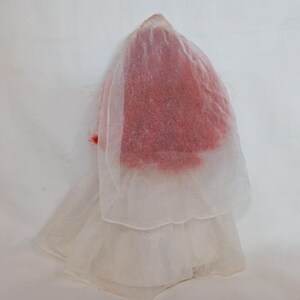 K&ouml;nnte beinhalten: Eine Vintage-Puppe in einem wei&szlig;en, mehrschichtigen Kleid mit einem durchsichtigen Schleier. Der Kopf der Puppe ist mit rotem Material bedeckt, und ein kleiner roter Akzent ist auf dem Schleier zu sehen. Die Puppe steht vor einem wei&szlig;en Hintergrund.
