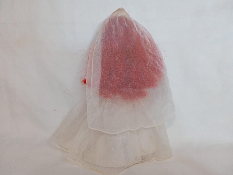 K&ouml;nnte beinhalten: Eine Vintage-Puppe in einem wei&szlig;en, mehrschichtigen Kleid mit einem durchsichtigen Schleier. Der Kopf der Puppe ist mit rotem Material bedeckt, und ein kleiner roter Akzent ist auf dem Schleier zu sehen. Die Puppe steht vor einem wei&szlig;en Hintergrund.