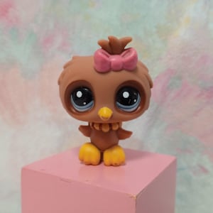 May include: A brown owl figurine with large, expressive eyes and a pink bow. The toy has yellow feet and a yellow beak. The owl is standing on a pink cube, with a colorful background.