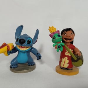 Puede incluir: Dos figuras de pl&aacute;stico azul y verde de Stitch y Lilo de la pel&iacute;cula de Disney Lilo & Stitch. Stitch sostiene una pistola de agua amarilla y Lilo sostiene una mu&ntilde;eca verde y un ukelele.