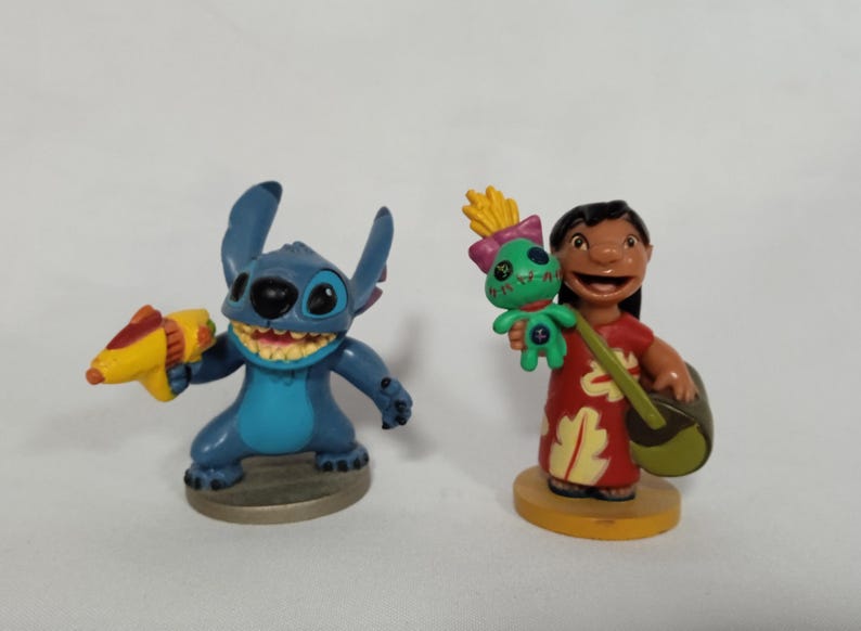 Puede incluir: Dos figuras de pl&aacute;stico azul y verde de Stitch y Lilo de la pel&iacute;cula de Disney Lilo & Stitch. Stitch sostiene una pistola de agua amarilla y Lilo sostiene una mu&ntilde;eca verde y un ukelele.