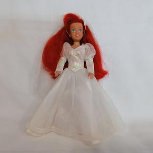 K&ouml;nnte beinhalten: Puppe einer Figur mit langen roten Haaren, die ein wei&szlig;es Hochzeitskleid mit langen &Auml;rmeln und einem Schleier tr&auml;gt. Die Puppe hat blaue Augen und einen l&auml;chelnden Gesichtsausdruck. Das Kleid hat ein paillettenbesetztes Oberteil und einen weiten Rock.