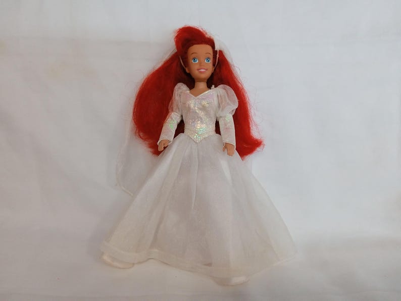 K&ouml;nnte beinhalten: Puppe einer Figur mit langen roten Haaren, die ein wei&szlig;es Hochzeitskleid mit langen &Auml;rmeln und einem Schleier tr&auml;gt. Die Puppe hat blaue Augen und einen l&auml;chelnden Gesichtsausdruck. Das Kleid hat ein paillettenbesetztes Oberteil und einen weiten Rock.
