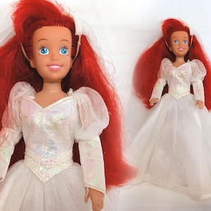 K&ouml;nnte beinhalten: Zwei Puppen einer Figur mit leuchtend roten Haaren und blauen Augen. Die Puppen tragen wei&szlig;e Hochzeitskleider mit langen &Auml;rmeln und einem T&uuml;llrock. Die Puppen haben einen wei&szlig;en Schleier und l&auml;cheln.