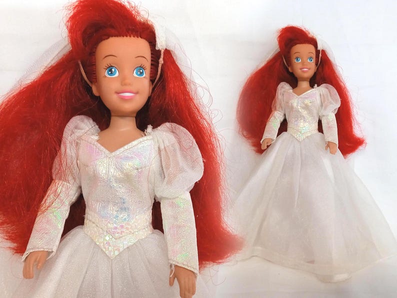 K&ouml;nnte beinhalten: Zwei Puppen einer Figur mit leuchtend roten Haaren und blauen Augen. Die Puppen tragen wei&szlig;e Hochzeitskleider mit langen &Auml;rmeln und einem T&uuml;llrock. Die Puppen haben einen wei&szlig;en Schleier und l&auml;cheln.