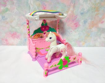 May include: A white toy pony with a pink and orange mane and tail stands in a pink bed with a canopy. The bed has a floral design and a rainbow above. The toy is a vintage collectible.