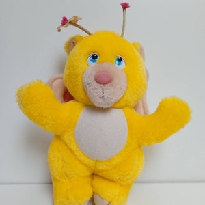 Könnte beinhalten: Ein gelber Plüschbär mit blauen Augen, einer rosa Nase und einem cremefarbenen Bauch. Der Bär hat kleine rosa Flügel und zwei Antennen mit rosa Blumenakzenten. Das Spielzeug steht mit ausgestreckten Armen da.