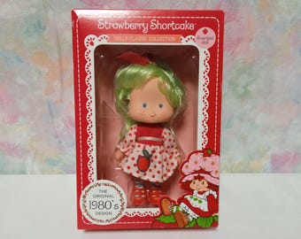 May include: A vintage Strawberry Shortcake doll in a red and white dress with a red bow in her hair. The doll is packaged in a red box with the words "Strawberry Shortcake Dolls Classic Collection" and "The Original 1980's Design" printed on it.