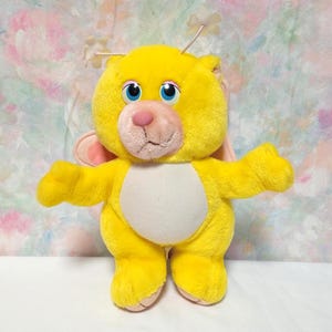 Könnte beinhalten: Ein gelbes Plüschtier, das einem Bären ähnelt, mit blauen Augen, einer rosa Nase und einem weißen Bauch. Es hat kleine rosa Ohren und antennenartige Merkmale. Das Spielzeug steht mit ausgestreckten Armen da.